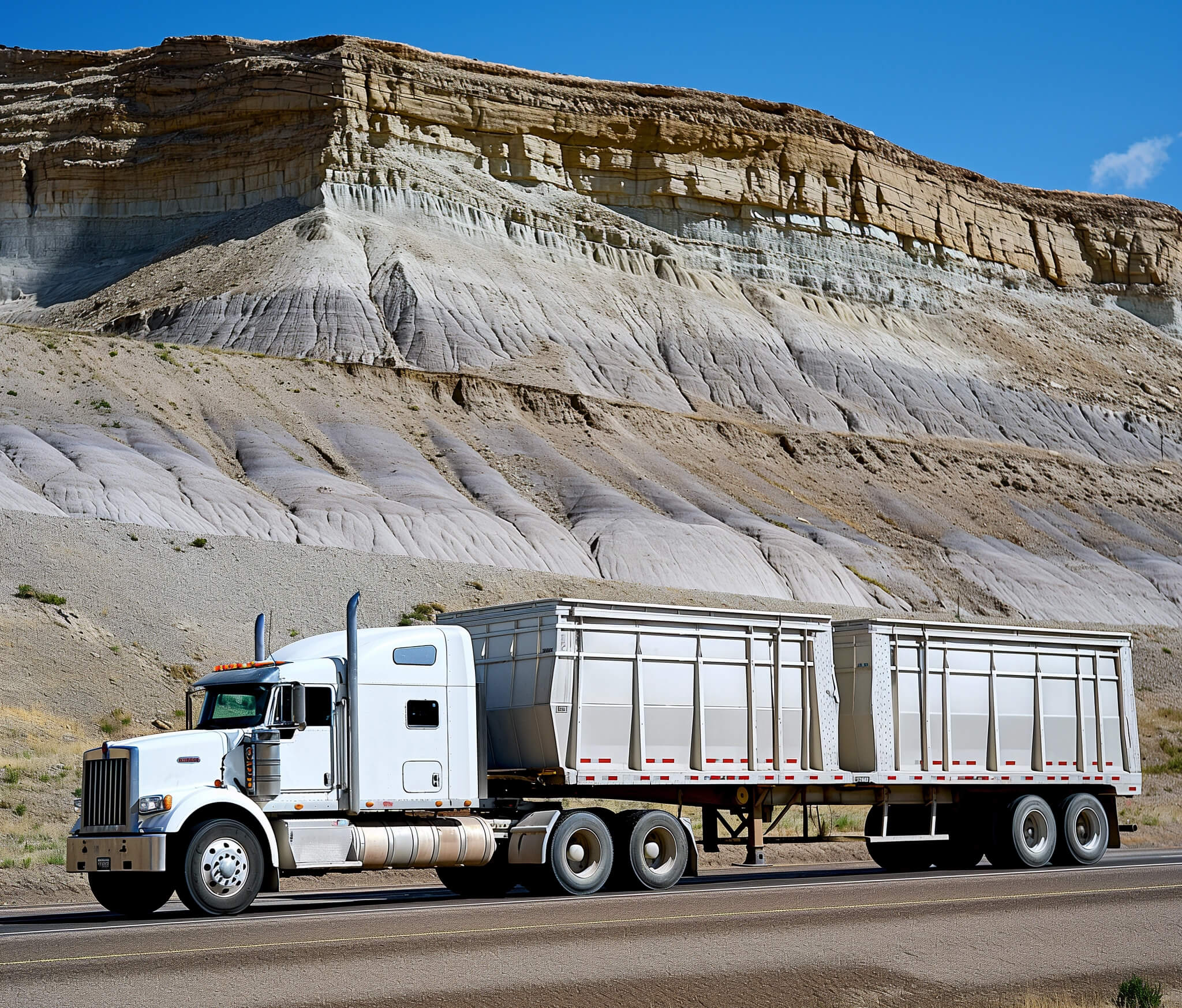 A white semi tractor fulling two trailers near a mountain